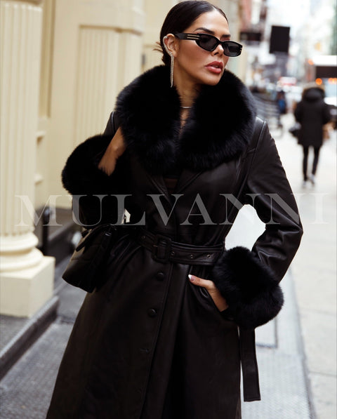 A woman wearing a sleek black leather trench coat featuring a large black fox fur collar and cuffs, exuding elegance and luxury. The rich textures of the leather and fur make this a bold statement piece.